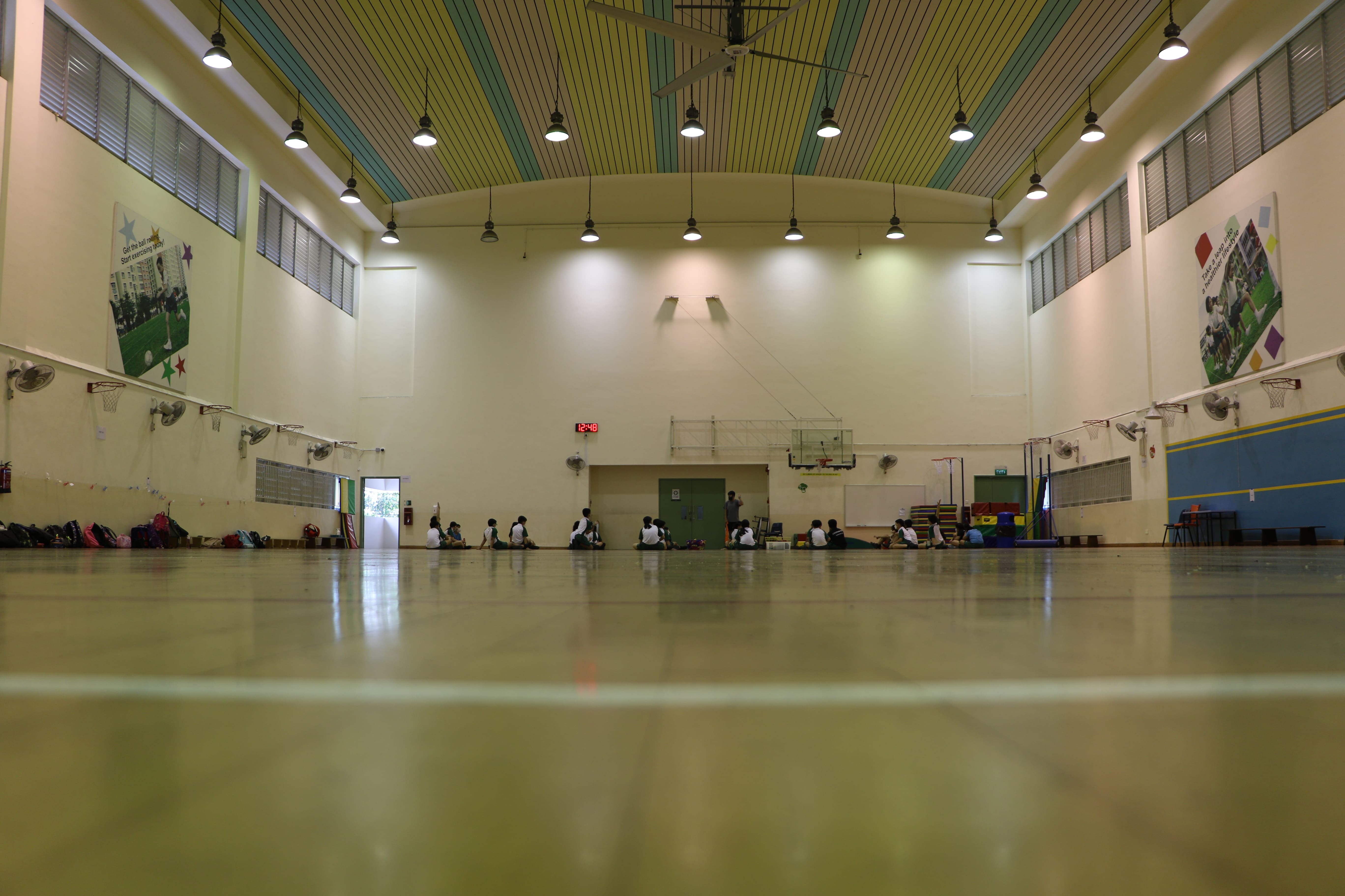 Spacious indoor sports hall with basketball hoops and students seated inside.